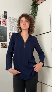 Joven mujer con cabello rizado y pendientes de aro, vestida con una camiseta de manga larga azul marino con cremallera, de pie en una habitación con arte en las paredes y plantas.