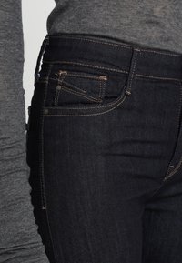 Tmavé džíny s kontrastním šitím, které mají standardní pětikapsový design a slim fit, který se přizpůsobuje postavě.