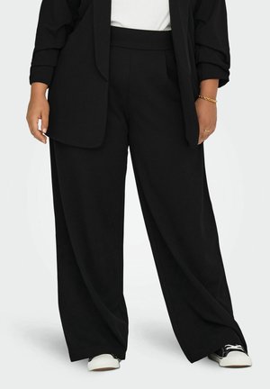 Kvinde iført sorte bukser med vide ben, sort blazer, hvid top og sorte og hvide sneakers, stående mod en ensfarvet baggrund.