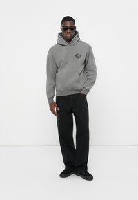 Grijze hoodie met een voorkantzak en logo, gecombineerd met zwarte wijde broeken en zwarte sneakers, gedragen door een persoon met zonnebril.