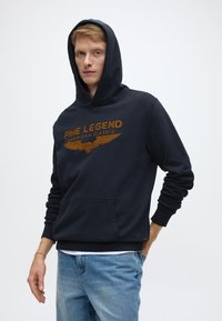 Temno modra jopa s kapuco in sprednjim žepom, z oranžnim napisom "PME LEGEND" in grafikonom kril. Gladek material in elastični manšeti.