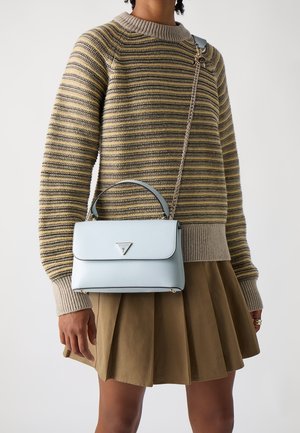 Person iført en stribet gul og grå sweater og en brun plisseret nederdel, der holder en lys blå håndtaske med kædesti.