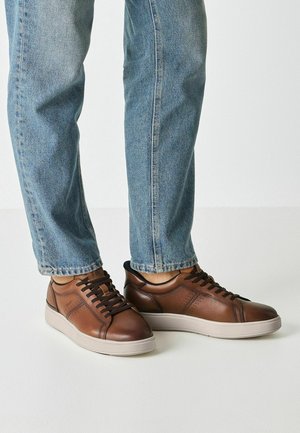 Brown leather sneakers featuring a smooth finish, perforated accents, and round toe on a light-colored rubber outsole, paired with blue jeans.