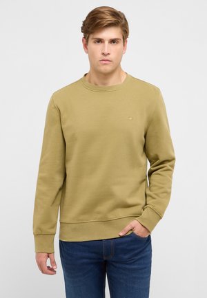 Junger Mann mit hellbraunen Haaren, der einen olivgrünen Pullover und blaue Jeans trägt, steht mit einer Hand in der Tasche vor einem einfarbigen Hintergrund.