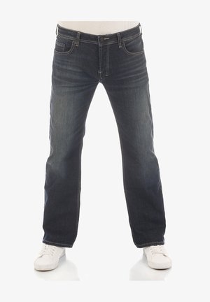 Jean en denim bleu foncé à coupe droite, coutures visibles et cinq poches. Porté avec des baskets blanches.