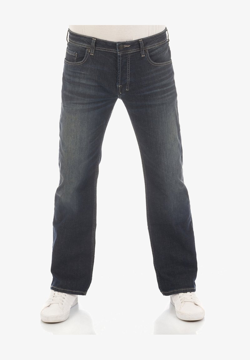 Jean en denim bleu foncé à coupe droite, coutures visibles et cinq poches. Porté avec des baskets blanches.