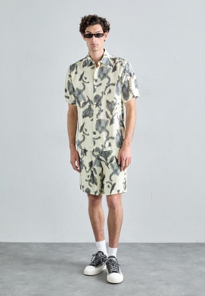 Short-sleeved, button-up shirt and matching shorts in light yellow with dark abstract patterns; paired with grey sneakers and black sunglasses.