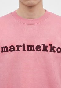Κοντινό πλάνο ατόμου που φοράει ροζ φούτερ με τη λέξη "marimekko" κεντημένη στο στήθος με σκούρα γράμματα.