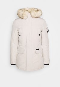Giacca imbottita beige chiaro con cappuccio in pelliccia sintetica, due tasche sul petto, tasche laterali e dettagli neri sui polsini e sull'orlo.