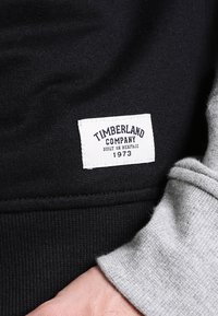 Zwarte en grijze sweater met een gestructureerde stof. Voorzien van een wit label met de tekst "TIMBERLAND COMPANY" en "1973" gedrukt in donkerblauw.