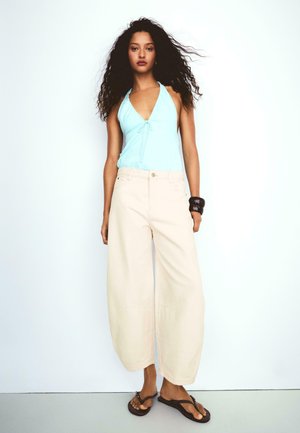 Kobieta z długimi kręconymi włosami, ubrana w jasnoniebieski top na szyję, szerokie beżowe spodnie, czarną bransoletkę i czarne klapki, stojąca na białym tle.