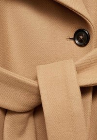 Béžový vlněný kabát s texturovaným vzorem tkaní, zdobený černým knoflíkem a zavazovacím páskem, který zdůrazňuje jeho pevnou konstrukci.