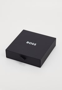 Svart låda med en slät matt yta, prydd med en silverfärgad "BOSS"-logotyp på toppen och en dragremsa för öppning. Fyrkantig form med skarpa hörn.