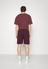 Homme portant un t-shirt bordeaux foncé et un short assorti, des chaussettes blanches et des baskets blanches, debout face à un mur gris clair uni.