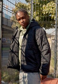 Sort, quiltet jakke med hætte, lagdelt over en lys grøn hoodie. Slidte denimjeans. Ude i naturen op ad et hegn med træer synlige.