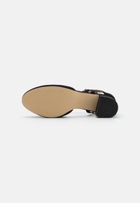 Chaussure à talons noire avec une tige texturée, un design ouvert et une semelle en caoutchouc beige. Dotée d'une sangle avec boucle pour un maintien sécurisé.
