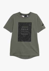 Olivi vihreä lyhythihainen t-paita, joka on valmistettu puuvillasta. Etupuolella on suuri musta kukkagraafi, jossa on "ADIDAS"-teksti useilla kielillä.