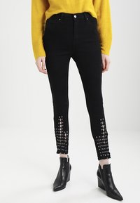 Černé skinny džíny z denimu s ozdobnými průhledy u kotníků. Vysoký pas; hladká textura; nošeny s žlutým svetr a černými kotníkovými botami.