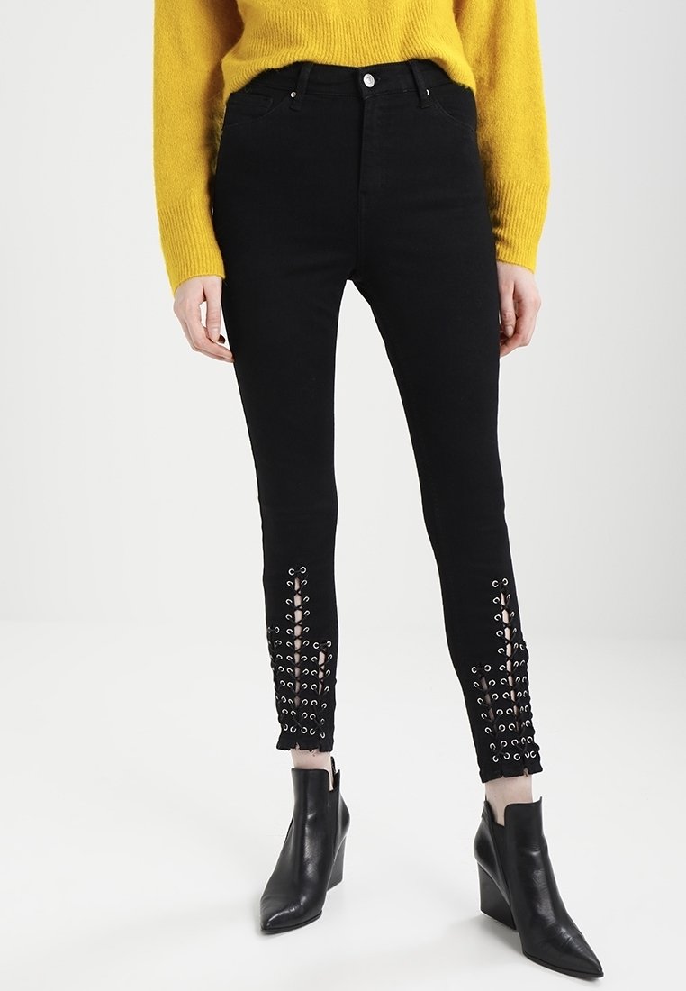 Černé skinny džíny z denimu s ozdobnými průhledy u kotníků. Vysoký pas; hladká textura; nošeny s žlutým svetr a černými kotníkovými botami.