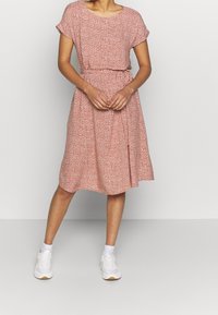 Mujer vestida con un vestido de estampado rosa a la altura de la rodilla, con mangas cortas y zapatillas blancas, de pie contra un fondo claro y uniforme.