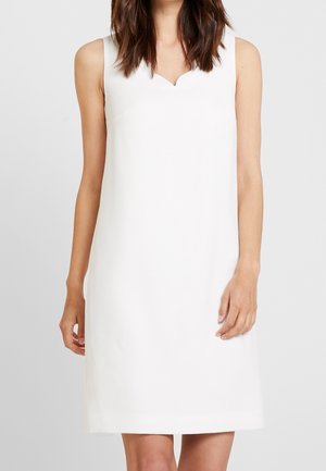 Robe blanche sans manches avec un col en V subtil portée par une personne aux cheveux bruns mi-longs sur un fond gris uni.