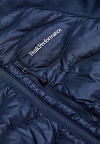 Marine geïsoleerde jas met een geplooide structuur, ritssluiting en het geborduurde 'Peak Performance' logo in het wit. Zacht, lichtgewicht materiaal.