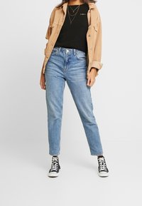 Brun manchesterjacka över en svart t-shirt, kombinerad med ljusblå högmidjade jeans och svarta högskor. Enkel lagerhalsband.