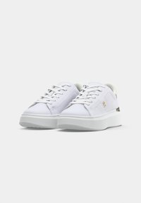 Un paio di sneakers bianche basse con suole spesse, design con lacci e piccolo logo dorato sul lato, su uno sfondo chiaro.