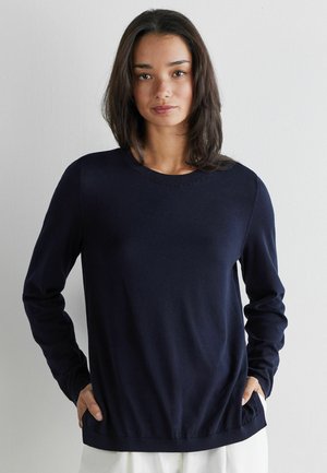 Ung kvinde med skulderlangt mørkt hår iført en marineblå langærmet sweater med hænderne i lommerne, stående mod en ensfarvet hvid baggrund.