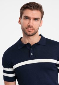 Mörkblått polo t-shirt med vita horisontella ränder, korta ärmar och en klassisk krage med två knappar. Slät stickad struktur.