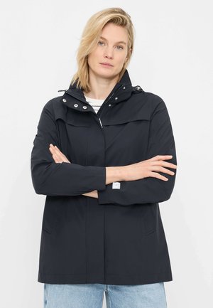 Woman with blonde hair wearing a black hooded jacket and light blue jeans, standing with arms crossed against a plain background.