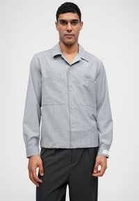 Chemise grise à boutons avec un col plat, deux poches poitrine et une texture subtile. Manches longues avec un détail de poignet contrasté.