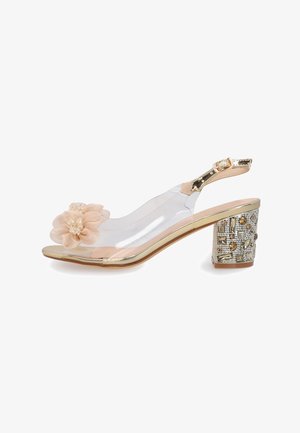 Clear heel sandal with gold trim, embellished block heel, transparent upper, and beige floral decoration on toe strap. Adjustable ankle strap.