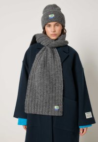 Naine, kes kannab tumesinist mantlit ja türkiissinist sviitrit, halli kootud salli kaelale mähitud ning halli kootud mütsi värviliste logodega.