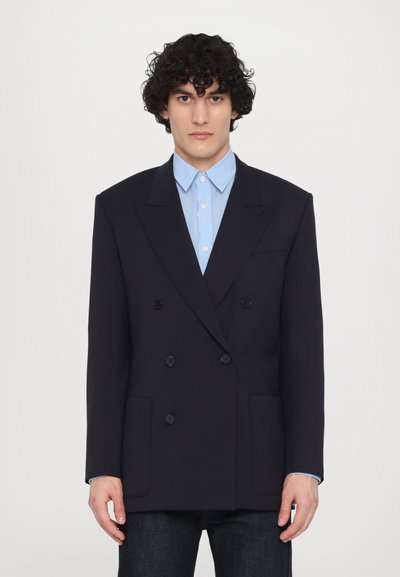 Blazer bleu marine à double boutonnage avec des revers crantés, deux poches frontales, boutons noirs, porté sur une chemise coll classique bleu clair, jeans droit.