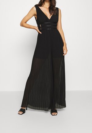 Femme portant une robe maxi plissée noire sans manches avec un décolleté en V profond et des sandales noires à talons sur fond blanc.