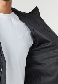 Sort lynlåsjakke med en glat tekstur, der har et hvidt logo på en hvid t-shirt under, hvilket fremhæver kontrasterende farver og materialer.