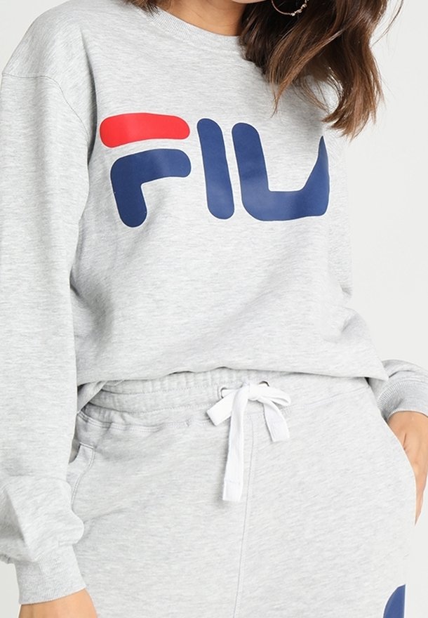 Femme portant un sweat-shirt FILA gris clair et un pantalon de survêtement assorti avec un cordon blanc au niveau du pantalon.