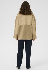 Keeruliste juustega inimene, kes kannab beežikat tuhksarnast jope ja tumedaid sirge säärega pükse, seisab seljaga vaataja poole lihtsa tausta ees.