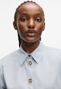 Ανοιχτό μπλε πουκάμισο με γιακά, εξοπλισμένο με χρυσά κουμπιά που φέρουν τα κουστούμια "K" και "A". Λείο υλικό, εφαρμογή ακριβώς στα μέτρα και καθαρός σχεδιασμός.