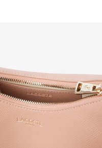 Pochette en cuir texturé rose avec une fermeture éclair dorée. Features logo Lacoste embossé à l'intérieur. Tirette de la fermeture éclair ornée de détails de logo.