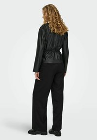 Musta keinonahkainen takki, jossa on vyötäröltä kiristettävä malli, pitkät hihat ja sileä pinta, yhdistetty mustiin leveälahkeisiin housuihin ja paksupohjaisiin mustiin kenkiin.