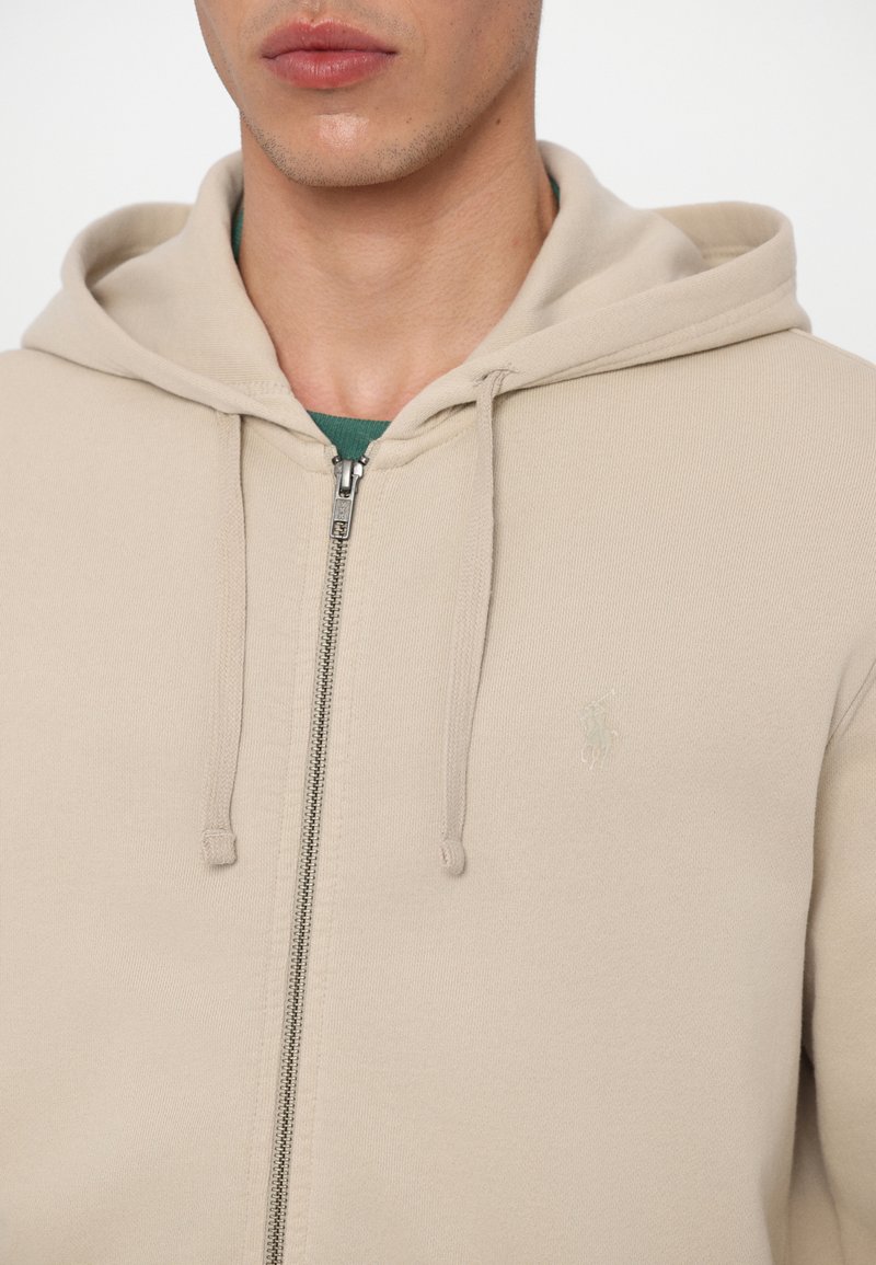 Beige zip-up hoodie met een zachte textuur, dubbele trekkoorden en een klein logo op de borst. Heeft een standaard pasvorm en capuchon.