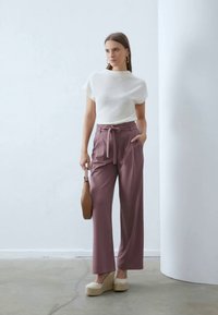Bílý top s krátkým rukávem, volný střih; růžové kalhoty s vysokým pasem a detailem na zavázání; béžové espadrilky na platformě; hnědá kabelka. Minmalistické pozadí.