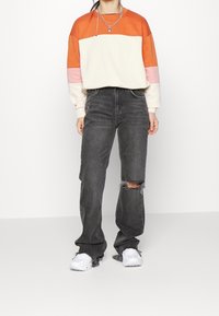 Osoba v mikině s barevnými bloky v oranžové, růžové a bílé barvě, roztrhaných černých džínách, bílých teniskách a stříbrném náhrdelníku na jednobarevném pozadí.