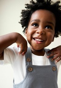 Copil mic zâmbitor cu păr creț, purtând o cămașă albă și o salopetă gri cu nasturi din lemn, ridicând mâinile aproape de umeri.