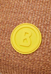 Geweven bruine oppervlakte met een gestructureerde afwerking, gekenmerkt door een opvallend rond geel rubberen embleem met de letter "B" in het midden.