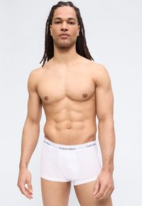 Μυώδης άνδρας με μακριά dreadlocks, φοράει λευκά μποξεράκια Calvin Klein, στέκεται μπροστά σε απλό φωτεινό φόντο.