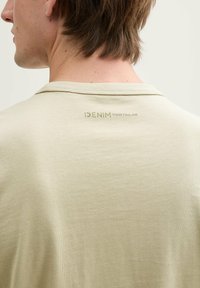 Világos zöld póló, puha tapintással, kerek nyakkal, a hátsó részén egy diszkrét "DENIM TOM TAILOR" nyomtatott logóval.