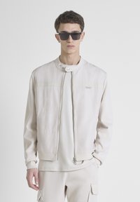 Chaqueta bomber de color beige claro con cuello alzado, cierre de cremallera y sutiles detalles de costura. Combinada con capas de color claro a juego por debajo.